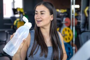 A person holds a clear water bottle, ready for a workout in a gym filled with weightlifting equipment and colorful plates in the background.