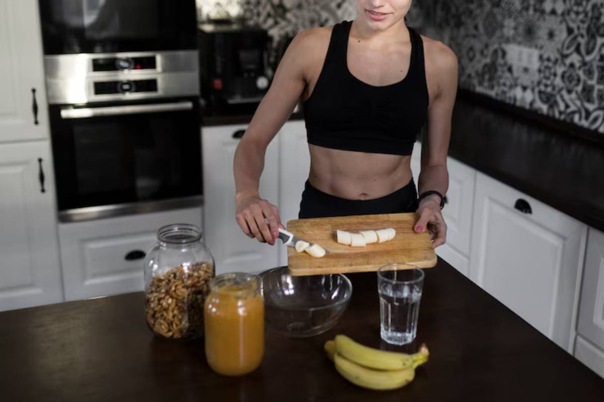 A fit woman in workout attire slices a banana on a wooden cutting board, with healthy snacks and drinks in a modern kitchen.
