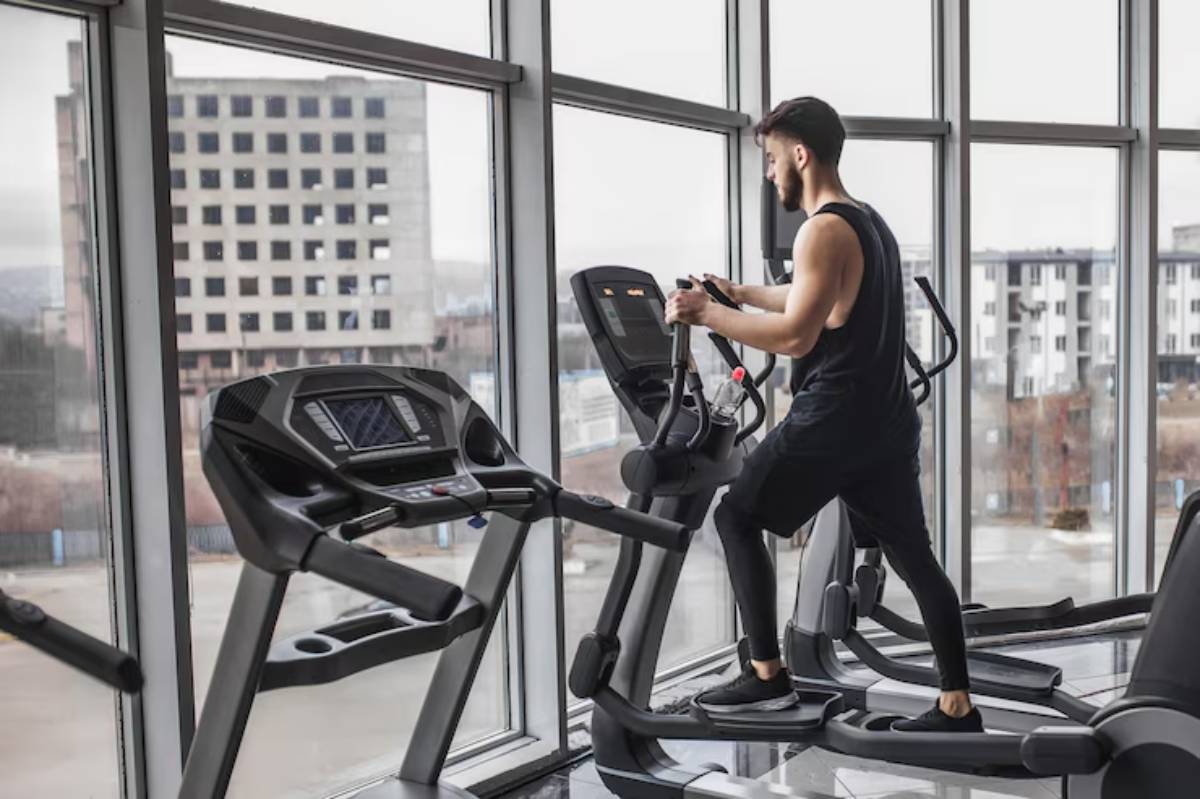 A person exercises on an elliptical machine in a modern gym with large windows, showcasing an urban landscape outside.