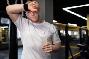 A person in a white shirt wipes sweat from their brow, holding a bottle of water with creatine in a gym setting.