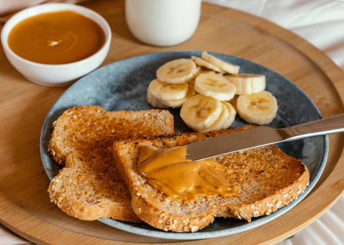 A plate with two slices of toasted whole grain bread spread with peanut butter, accompanied by banana slices and a small bowl of sauce.