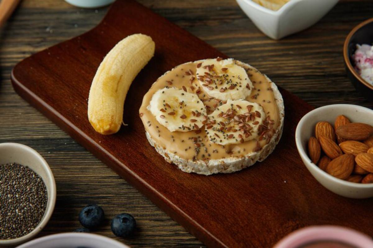 A rice cake topped with peanut butter, banana slices, flaxseeds, and chia seeds, next to a banana and various nuts on a wooden board.