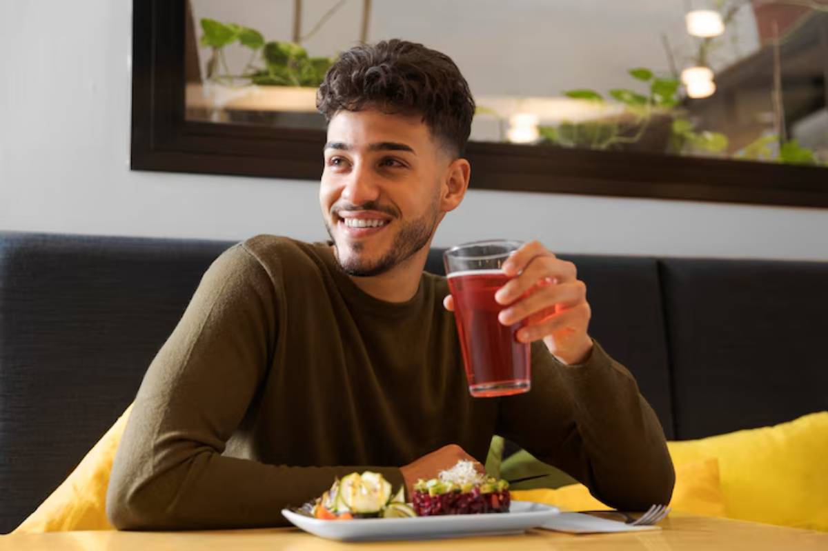A person holds a glass of red drink while sitting at a table with a plate of colorful sushi and garnished vegetables.