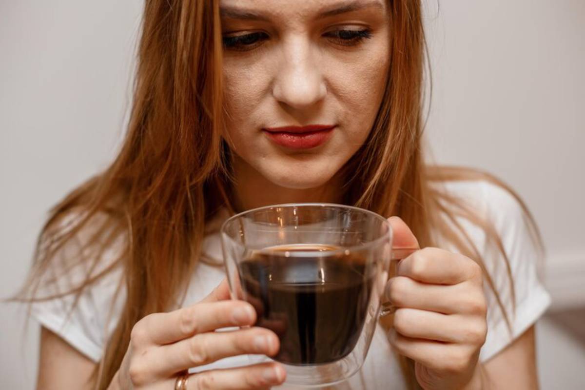 A person holds a transparent cup filled with dark coffee, with long brown hair cascading over a white shirt in a neutral background.