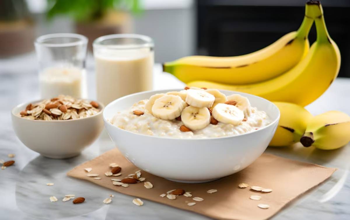 A bowl of oatmeal topped with banana slices and almonds, with bananas and glasses of milk in the background. Healthy breakfast scene.