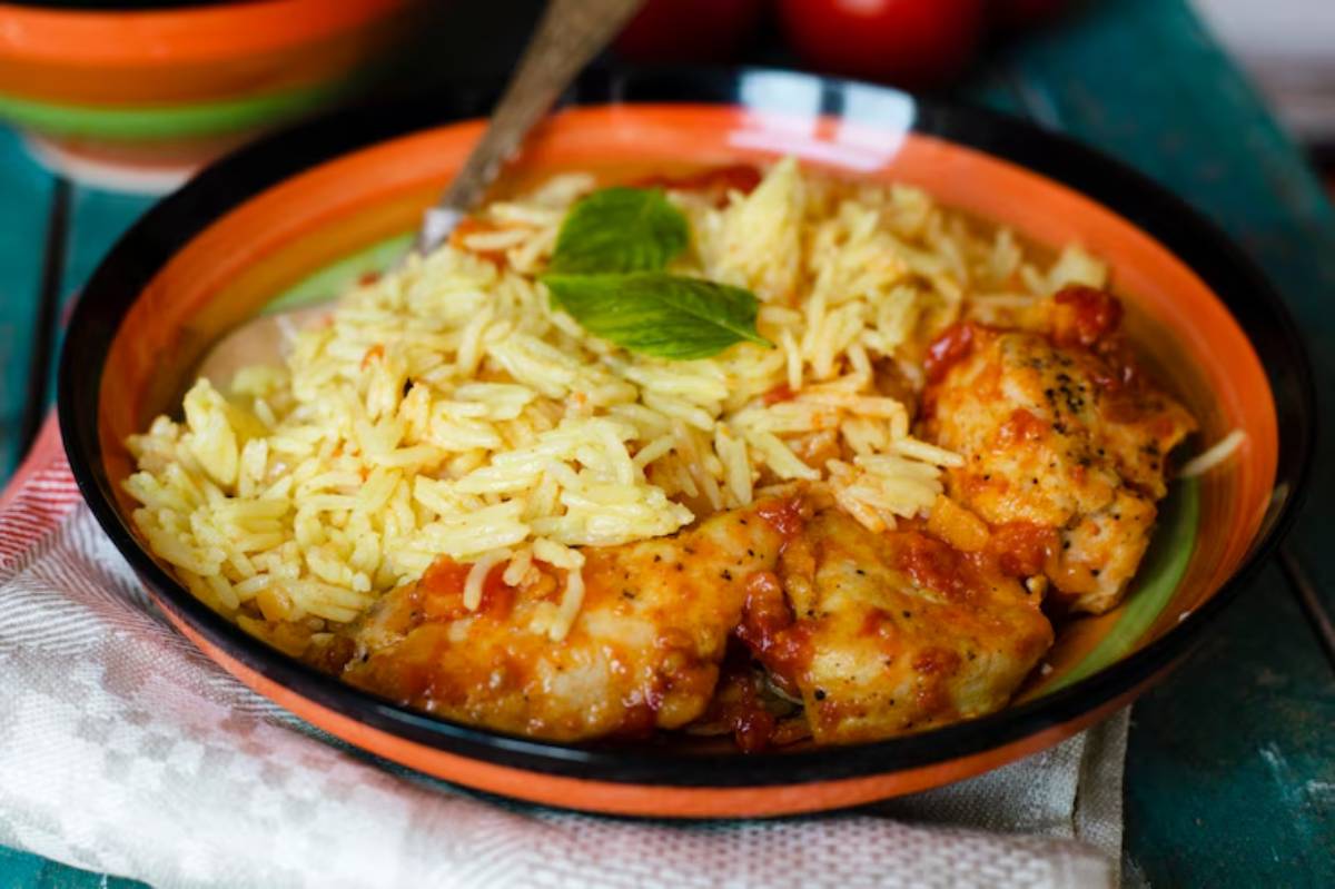 A vibrant bowl of chicken cooked in tomato sauce served with fluffy yellow rice, garnished with a fresh basil leaf.