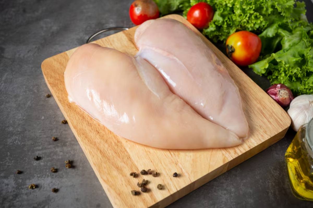 Two raw chicken breasts on a wooden cutting board, surrounded by fresh vegetables and spices, on a dark kitchen surface.