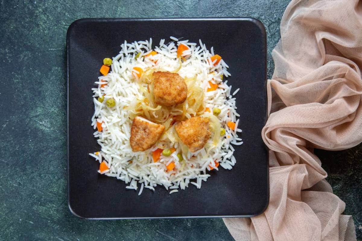 A black plate holds fluffy white rice topped with sautéed onions, colorful diced vegetables, and three golden-brown breaded pieces.