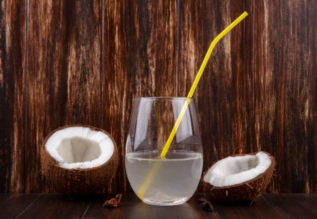 A glass of coconut water with a yellow straw, surrounded by two halved coconuts on a wooden table.