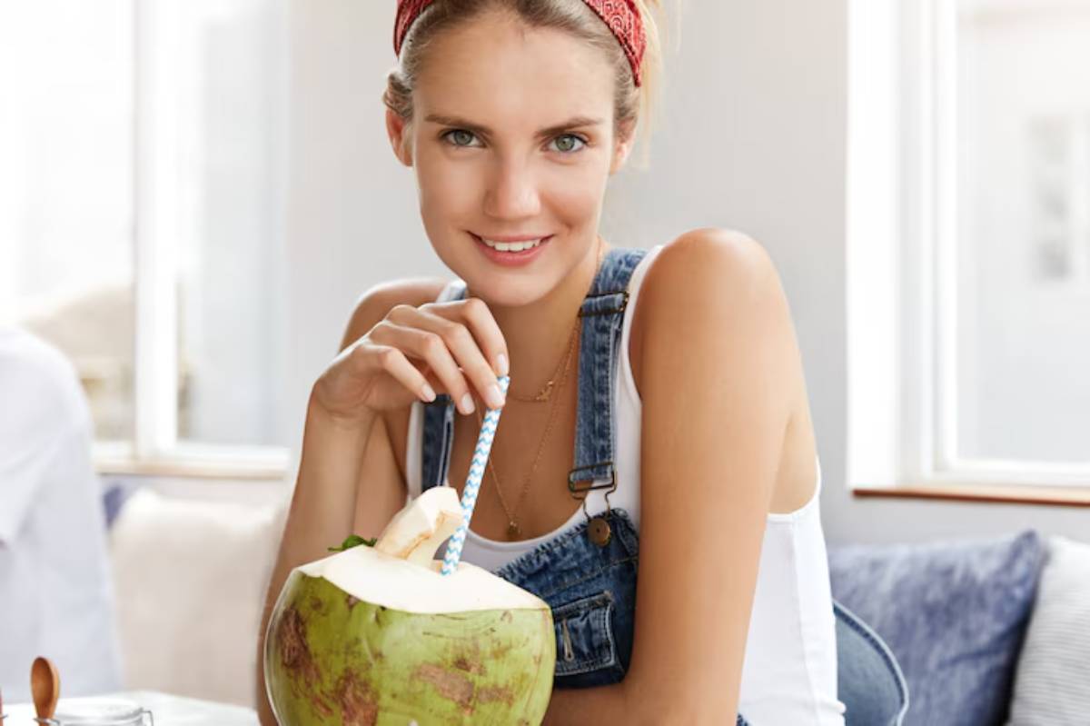 A young woman in denim overalls sips from a coconut with a straw, enjoying a refreshing drink in a sunlit café.