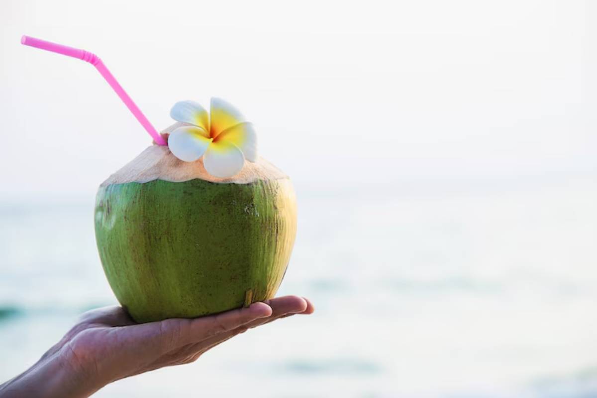 A hand holding a coconut drink with a pink straw and a yellow flower, against a blurred beach background.