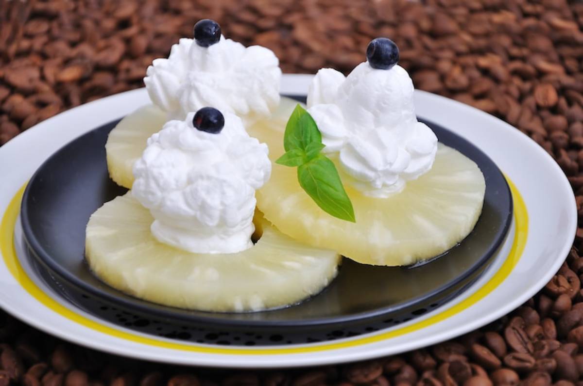 Three pineapple slices topped with whipped cream and blueberries, garnished with a fresh basil leaf, on a black plate.