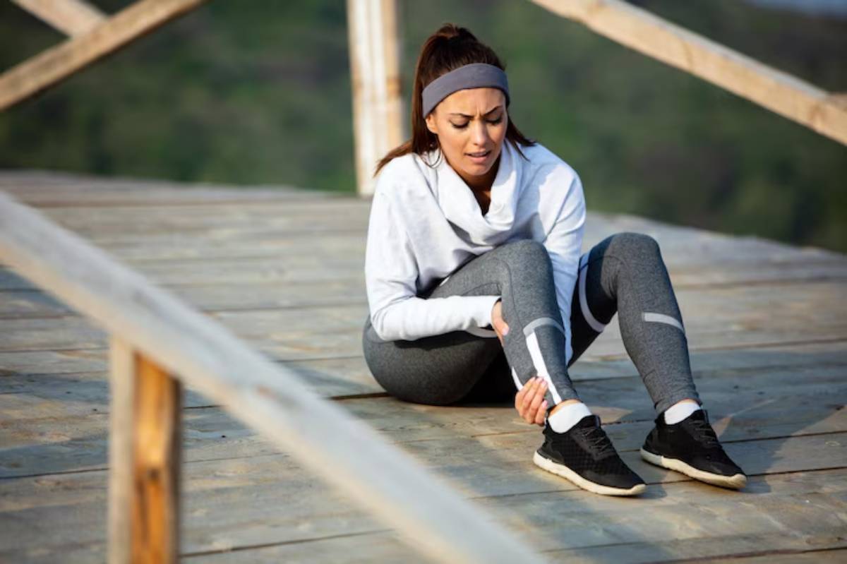 A person in athletic wear sits on a wooden deck, holding their ankle, surrounded by a lush green landscape.