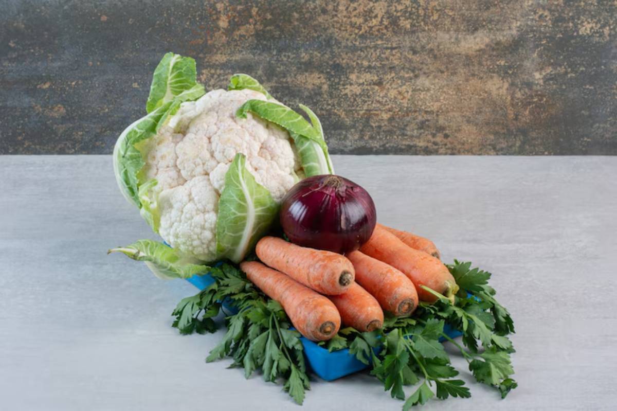 A vibrant display of fresh vegetables: a head of cauliflower, red onion, carrots, and parsley on a light background.
