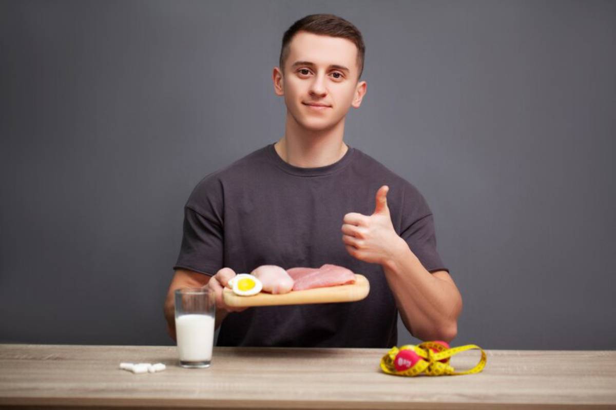 A person gives a thumbs-up while holding a wooden platter with raw meat and a boiled egg, alongside a glass of milk and a measuring tape.