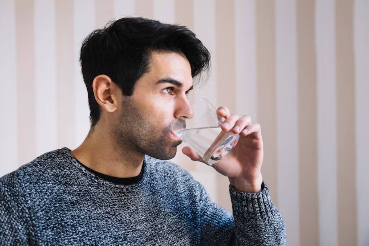 A person in a gray sweater is sipping water from a clear glass, against a light striped wall backdrop.