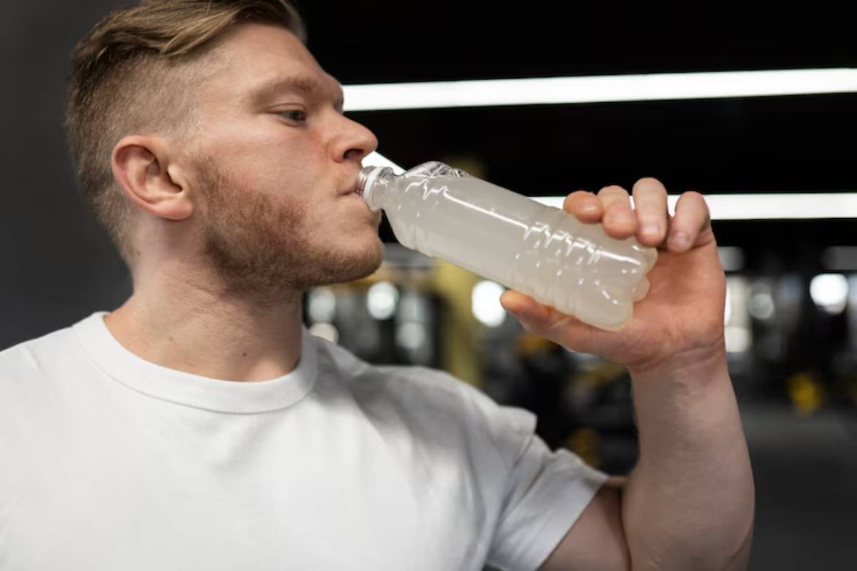 A person wearing a white t-shirt drinks from a transparent water bottle in a gym setting, with equipment blurred in the background.