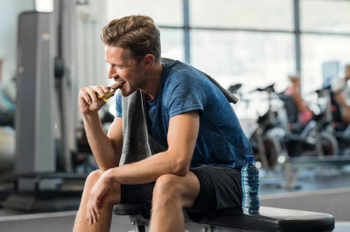 A young man in a blue athletic shirt sits on a bench in a gym, holding a snack and a water bottle, with workout equipment in the background.