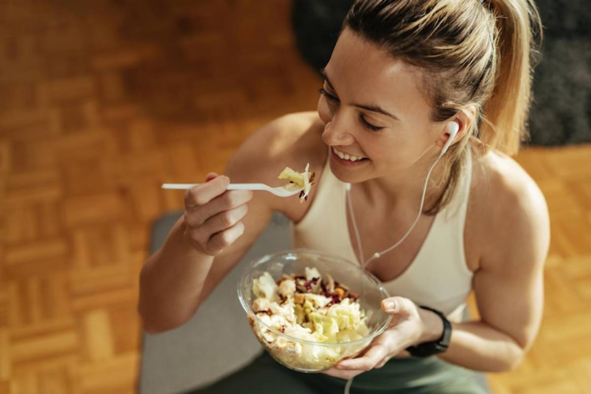 A fitness enthusiast enjoys a colorful salad in a stylish bowl, wearing headphones, seated on a wooden floor.