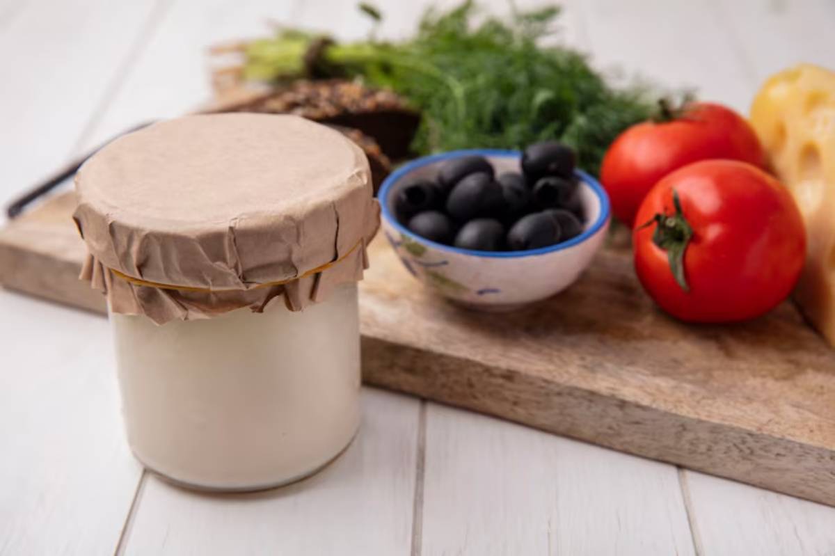 A glass jar of yogurt with a paper lid sits on a wooden board, surrounded by fresh tomatoes, olives, cheese, and green herbs.