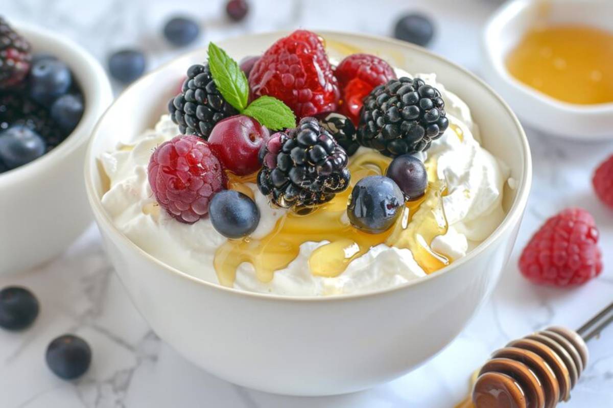 A bowl of whipped cream topped with mixed berries, drizzled with honey, and garnished with mint leaves, surrounded by more berries.