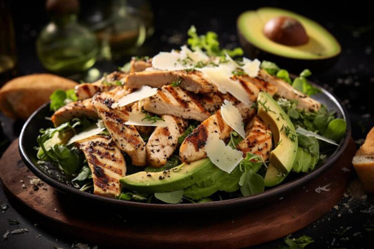 A vibrant salad featuring grilled chicken, avocado slices, greens, and shaved Parmesan, served in a dark, rustic setting.
