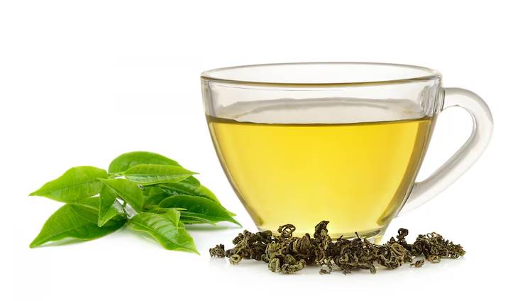 A clear glass cup filled with pale green tea, surrounded by green tea leaves and dried tea leaves on a white background.