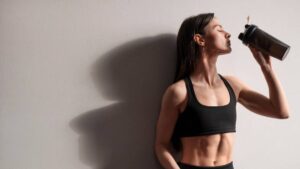 A fit person in a black crop top drinks from a shaker bottle, leaning casually against a light-colored wall.