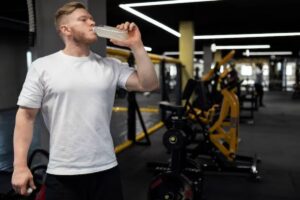 A person in a white t-shirt drinks from a sports bottle inside a modern gym, surrounded by workout equipment and bright lighting.