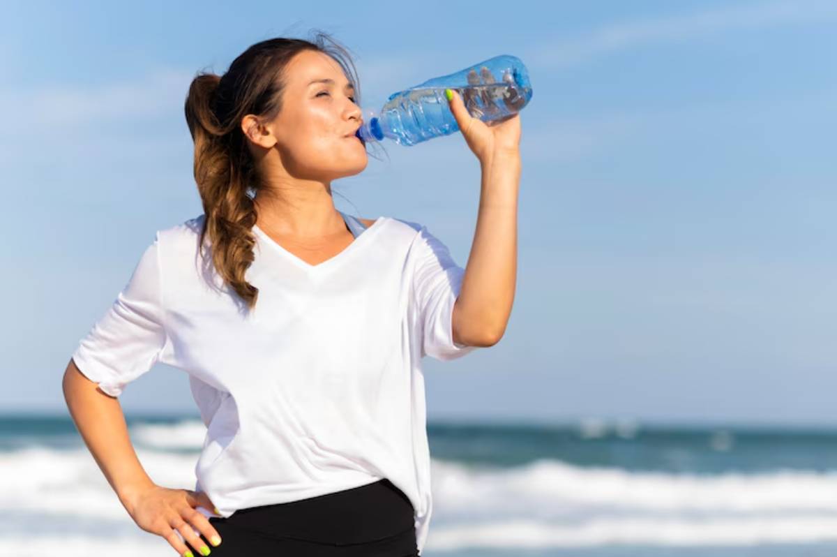 A woman in a white shirt drinks water from a plastic bottle, standing on a beach with waves and a clear blue sky in the background.