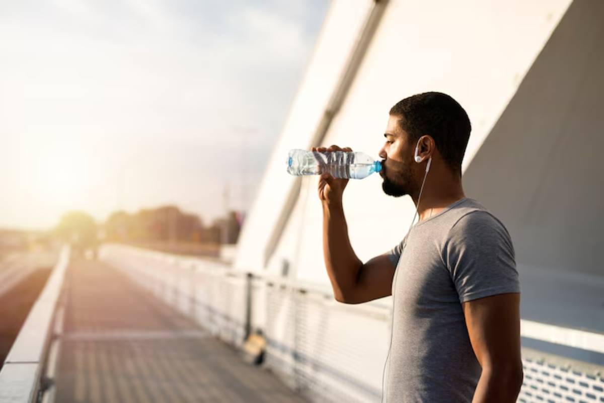 A person in a gray t-shirt drinks water while listening to music on earbuds, standing on a sunlit bridge.