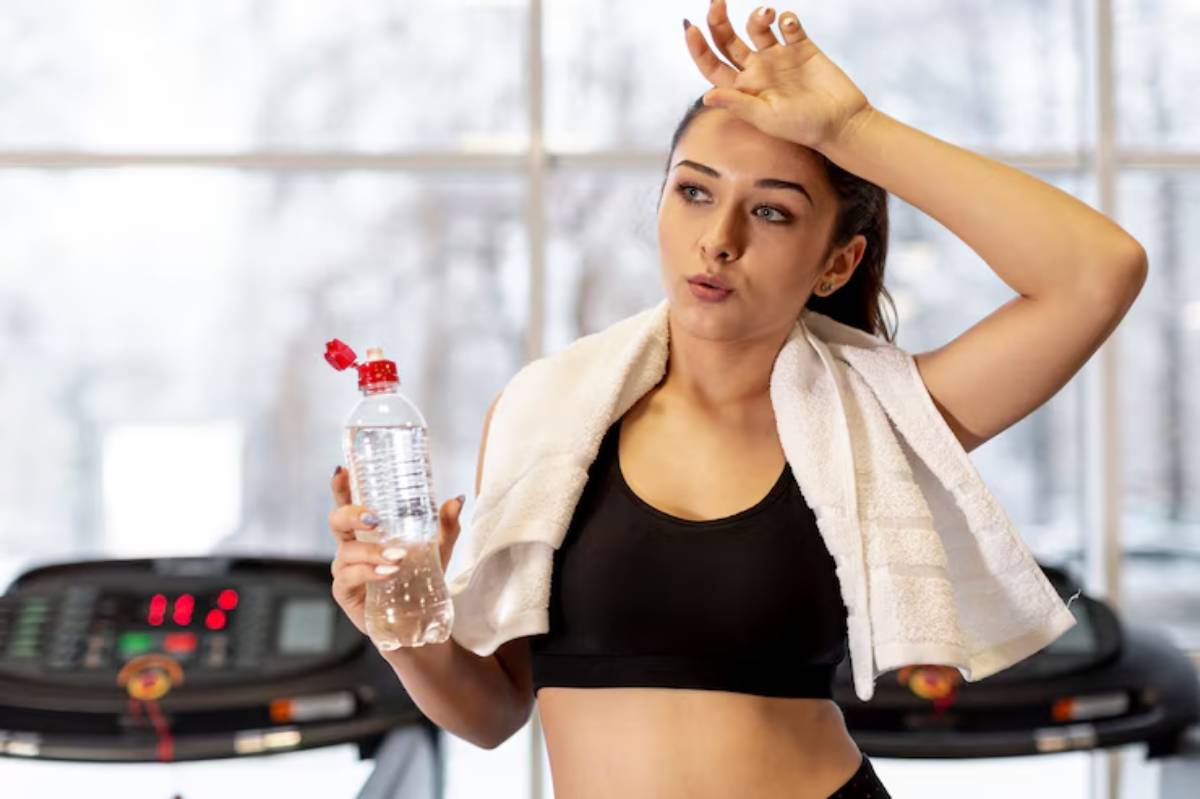 A woman in workout attire holds a water bottle and wipes her forehead with a towel, surrounded by gym equipment.