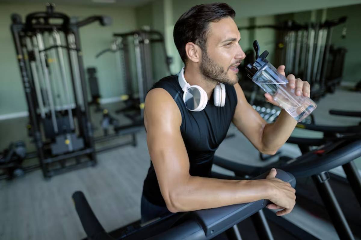 A fit individual in a sleeveless top and headphones takes a break while holding a water bottle in a gym setting.