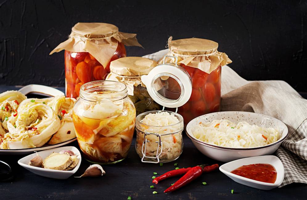 Various jars of pickled vegetables and kimchi, alongside fresh cabbage, garlic, ginger, chili peppers, and dipping sauce on a dark background.