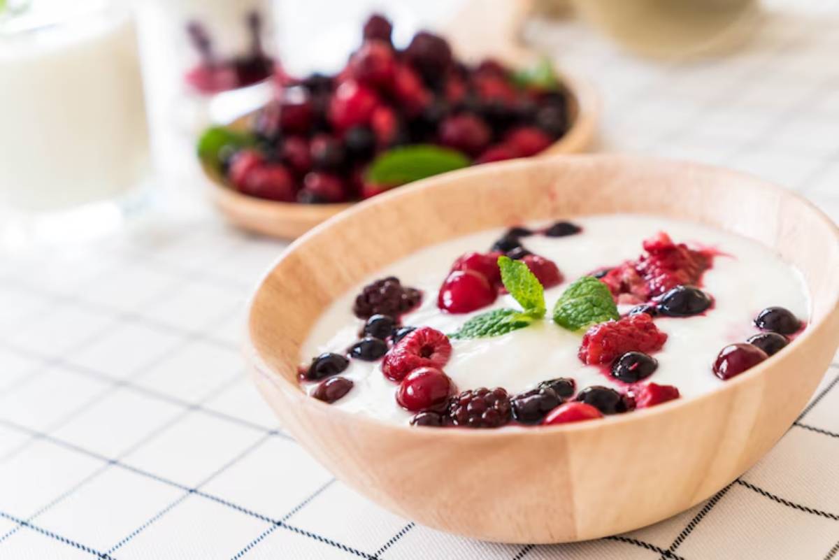 A wooden bowl filled with yogurt topped with fresh mixed berries and mint leaves, with more berries in a bowl in the background on a checkered tablecloth.
