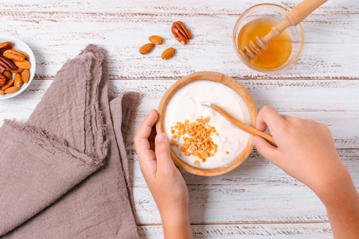 A person stirring yogurt with granola in a wooden bowl, surrounded by almonds, pecans, honey, and a textured cloth on a wooden surface.