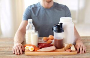A man sits with a variety of protein-rich foods, including eggs, milk, fish, chicken, and protein powder, on a wooden table.