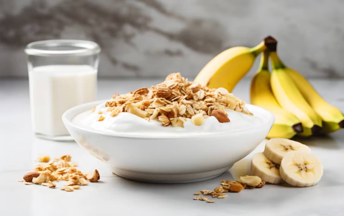 A bowl of yogurt topped with granola and sliced bananas, alongside a glass of milk and whole bananas in the background.