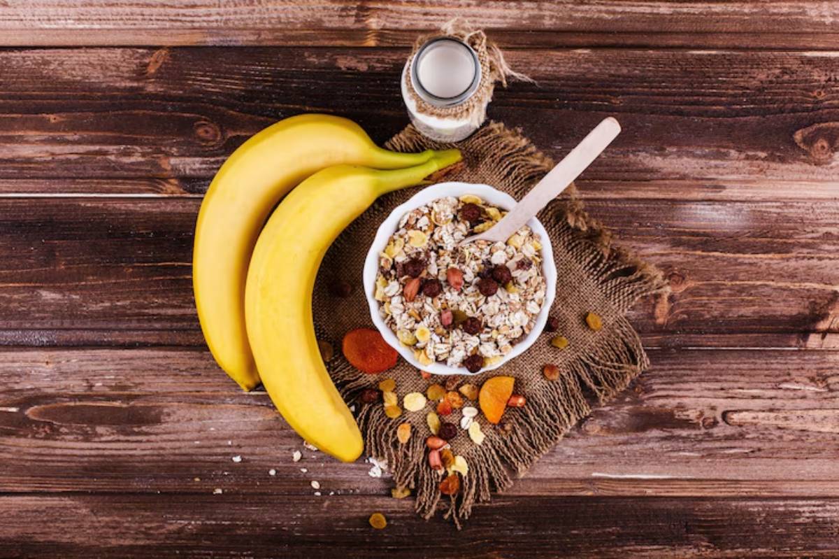A bowl of granola with dried fruits and nuts, two ripe bananas, and a jar of milk on a burlap mat over a wooden surface.