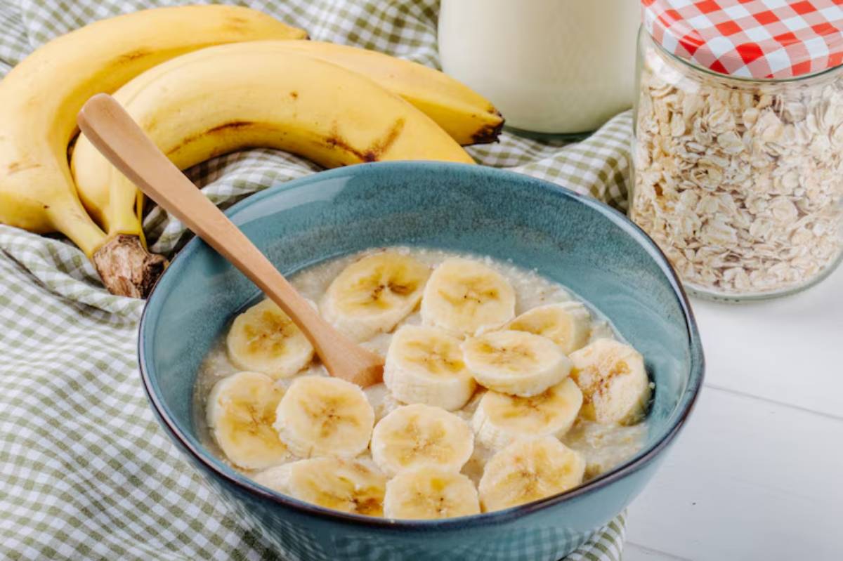 A blue bowl filled with oatmeal topped with banana slices, accompanied by bananas, a jar of oats, and milk on a checkered tablecloth.