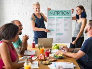 A group of people engages in a nutrition workshop, examining a poster listing food categories like carbs, proteins, and fats, with colorful drinks.