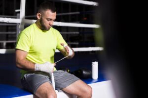 A boxer in a bright yellow shirt wraps his hands, preparing for training in a gym surrounded by boxing equipment.