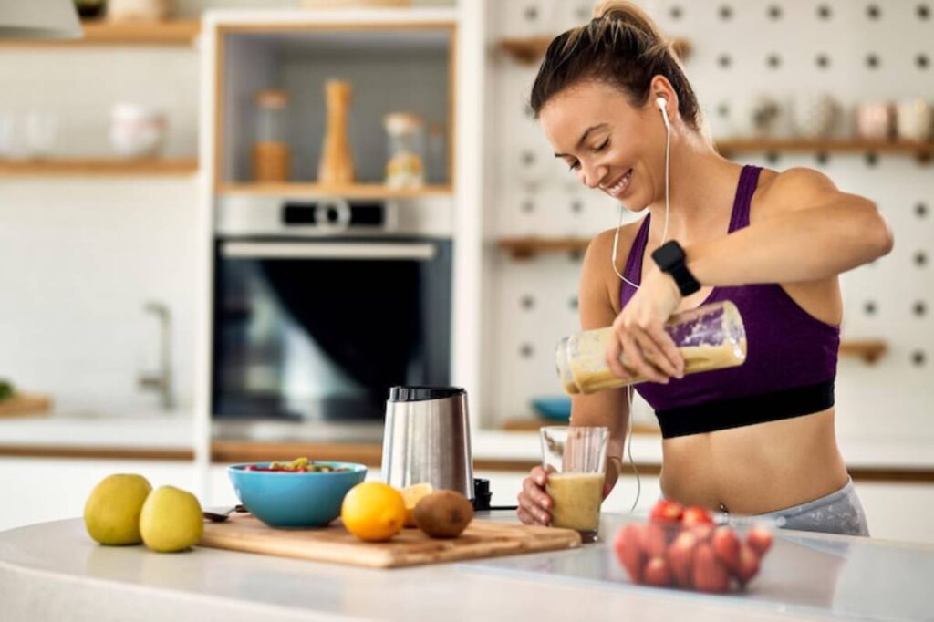 A woman in a purple sports bra prepares a smoothie in a modern kitchen, surrounded by fruits and a bowl of cereal.
