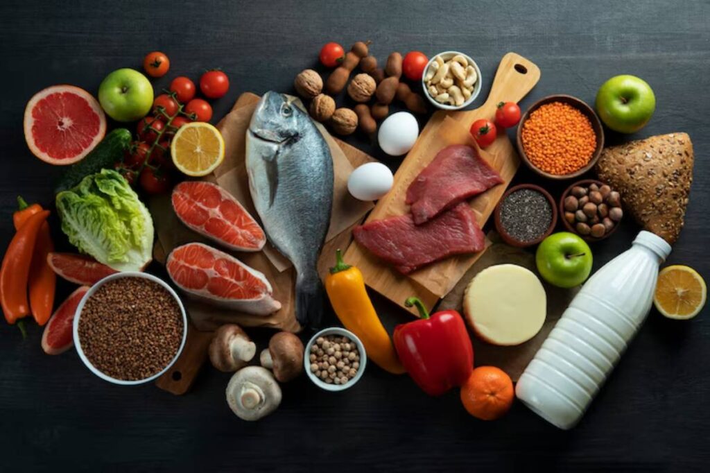 A variety of fresh foods including fish, meats, fruits, vegetables, nuts, and dairy arranged on a dark wooden surface.