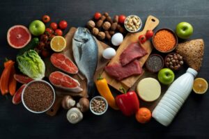 A variety of fresh foods including fish, meats, fruits, vegetables, nuts, and dairy arranged on a dark wooden surface.