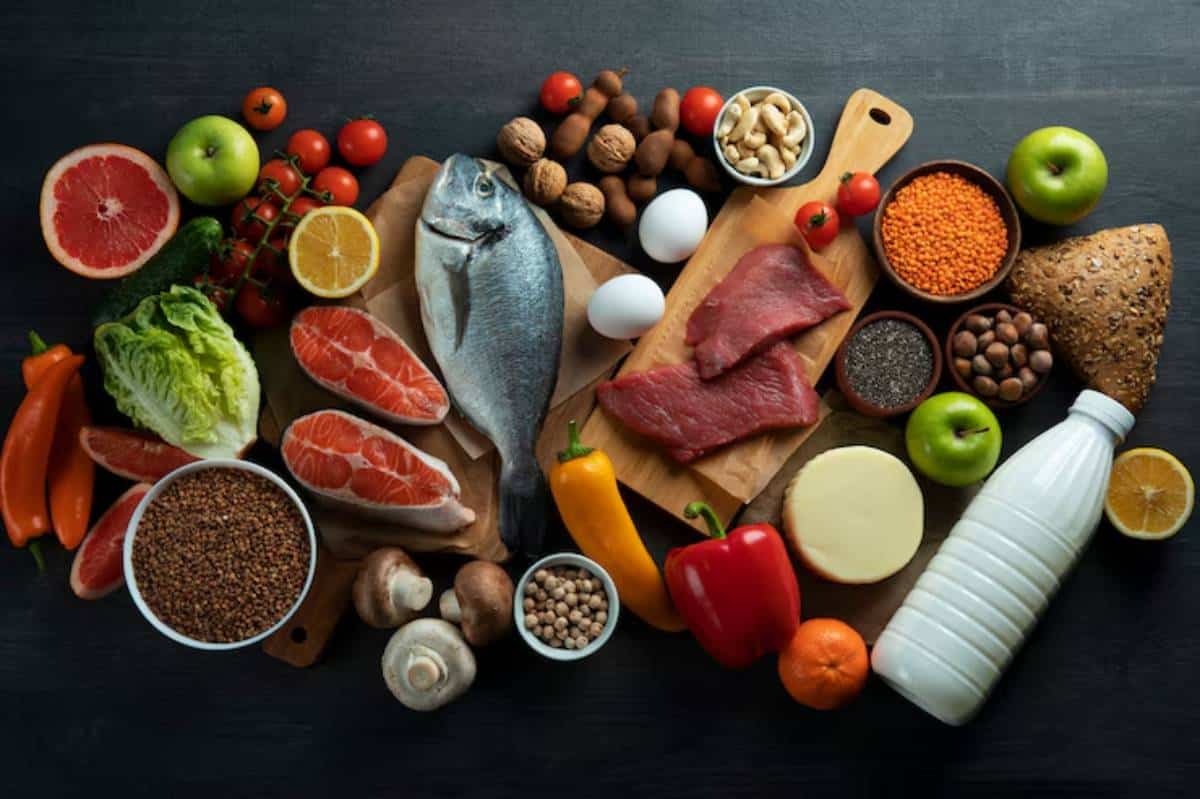 A variety of fresh foods including fish, meats, fruits, vegetables, nuts, and dairy arranged on a dark wooden surface.
