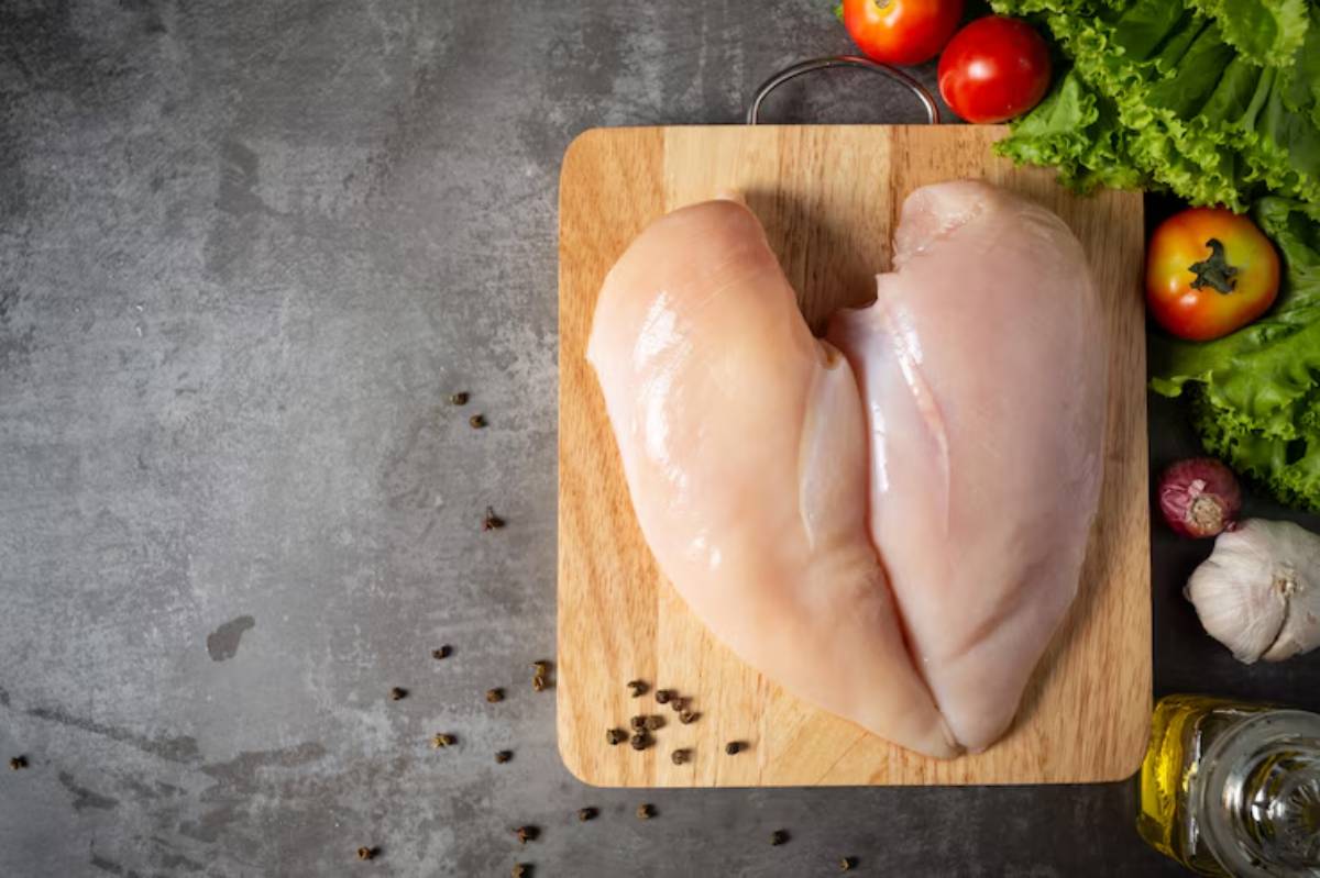 Raw chicken breast on a wooden cutting board, surrounded by fresh tomatoes, lettuce, garlic, and oil on a gray countertop.