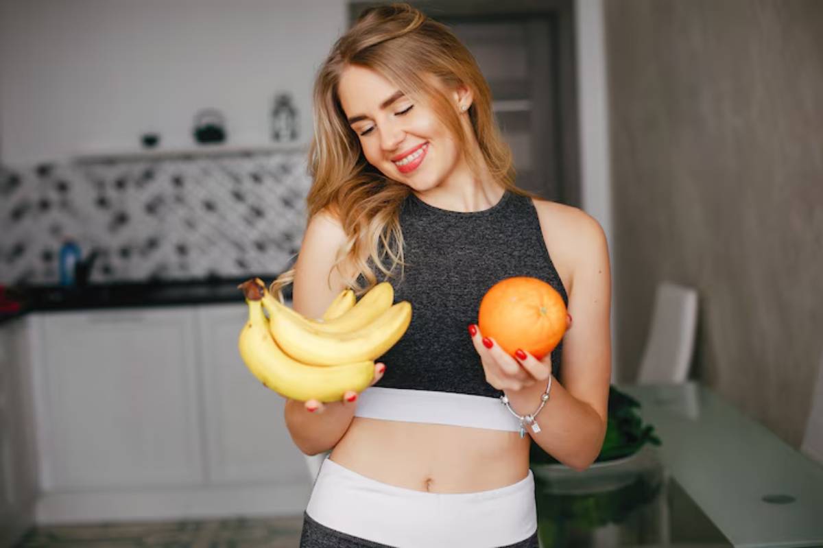 A woman in a sports outfit holds a bunch of bananas in one hand and an orange in the other, standing in a modern kitchen.