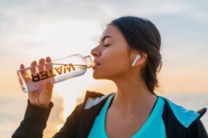 A woman drinks from a clear bottle labeled "WATER," wearing wireless earbuds, against a sunset at the beach.