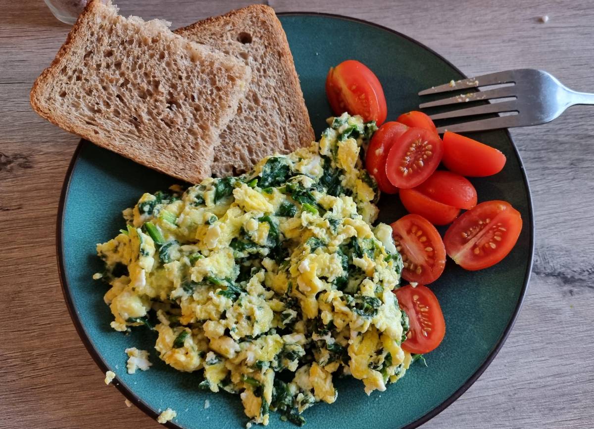 A plate of scrambled eggs with spinach, cherry tomatoes, and two slices of whole grain bread, set on a wooden table.
